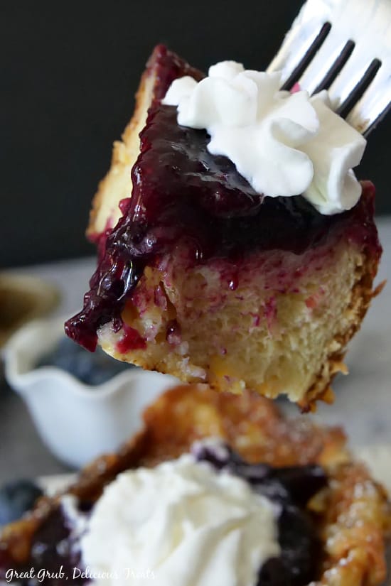French Toast with Blueberry Topping is a thick, delicious French toast using Challah bread dipped in a delicious eggy mixture, fried then baked, topped with blueberries and whipped cream. A close up photo of a bite of French Toast with Blueberry Topping on a fork.