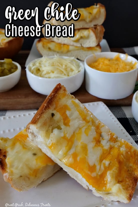 A white plate with two slices of green chile cheese bread on it with more slices in the background along with more shredded cheese and diced green chilies in small white bowls.