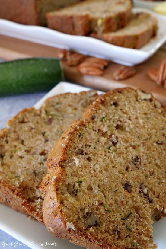 Zucchini Bread - two slices placed on a white plate with pecans, zucchini and more slices of zucchini bread in the background.