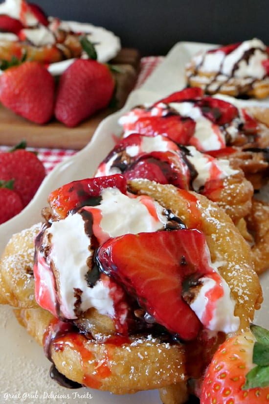 A close up picture of a Funnel Cake on a white plate, topped with powdered sugar, fresh strawberries, whipped cream, and drizzled with chocolate syrup. 