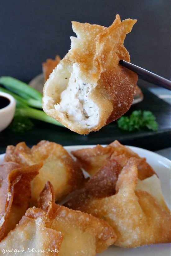 A single wonton being held up with chopsticks, with a bite taken out showing the cream cheese filling, over a white plate with more cream cheese wontons below.