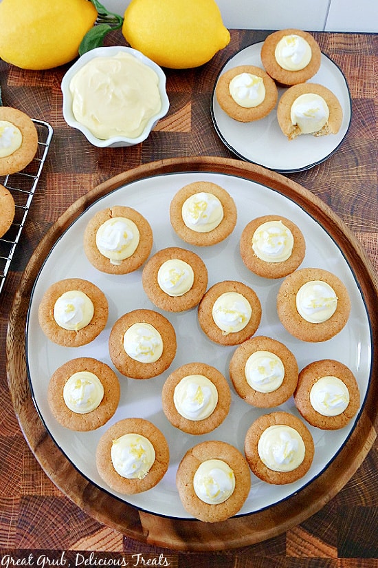 A white plate with 16 mini lemonade pie cookie bites on it and another small plate with 3 cookie bites and a small bowl filled with lemonade cream cheese pie filling and two lemons in the background.