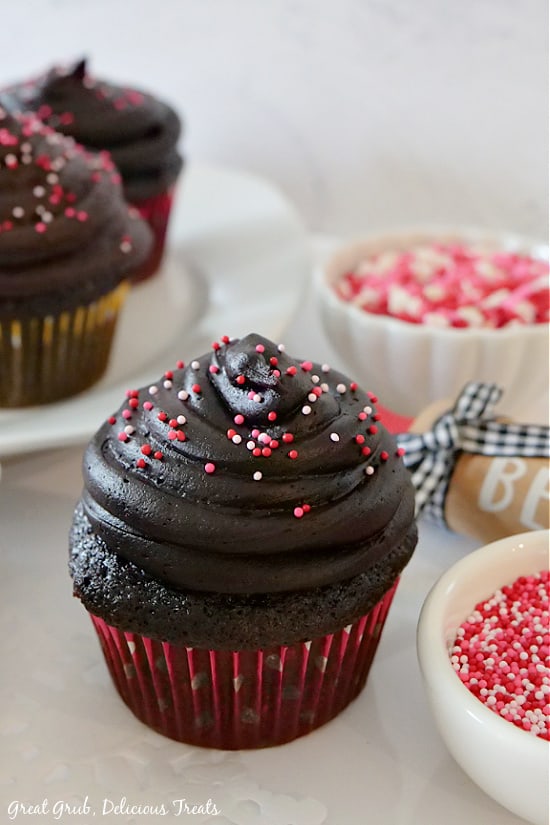 A single dark chocolate zucchini cupcake with dark chocolate frosting and sprinkles with two more cupcakes and two bowls of sprinkles in the background.