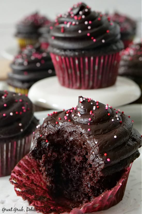 A dark chocolate zucchini cupcake with a bite taken out of it sitting on a white plate with more cupcakes in the background.