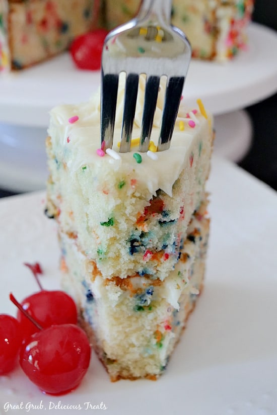 A slice of funfetti cake with a fork placed in the top of the slice and 3 cherries placed next to the slice of cake.