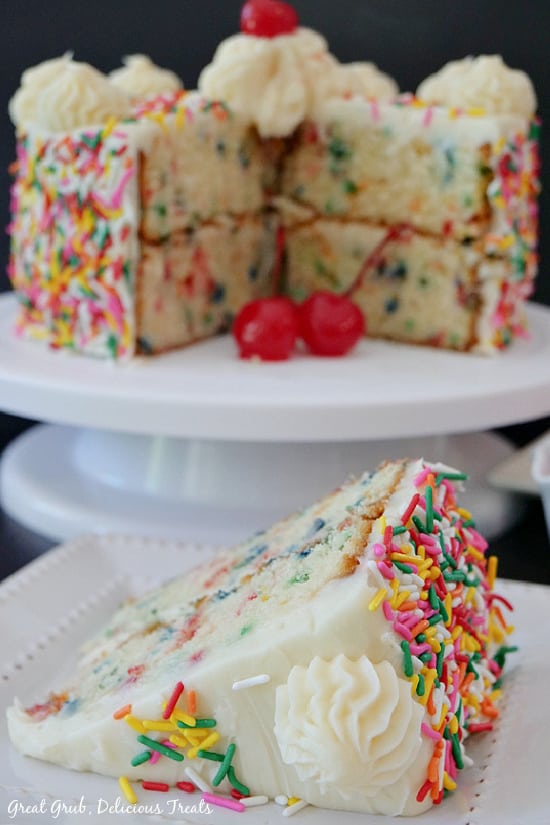 A funfetti cake sitting on a white cake stand with a piece taken out and placed on a white plate.
