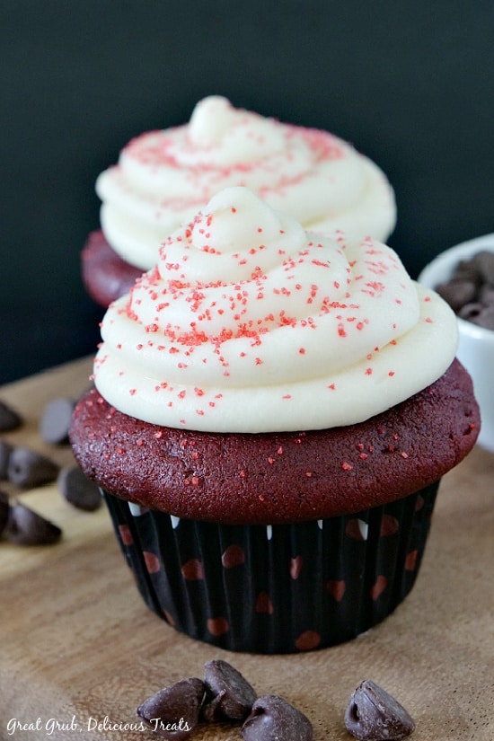 Two red velvet cupcakes with cream cheese frosting and sprinkles on it placed on a wood cutting board with chocolate chips laying on the board.