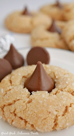 A single peanut butter blossom cookies on a white plate with 3 chocolate kisses next to it, with more cookies in the background.