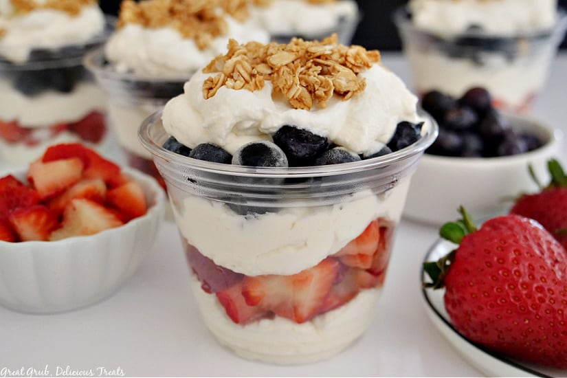 Small white bowls with whole strawberries and blueberries a clear container layered with whipped cream, strawberries, blueberries, and granola.