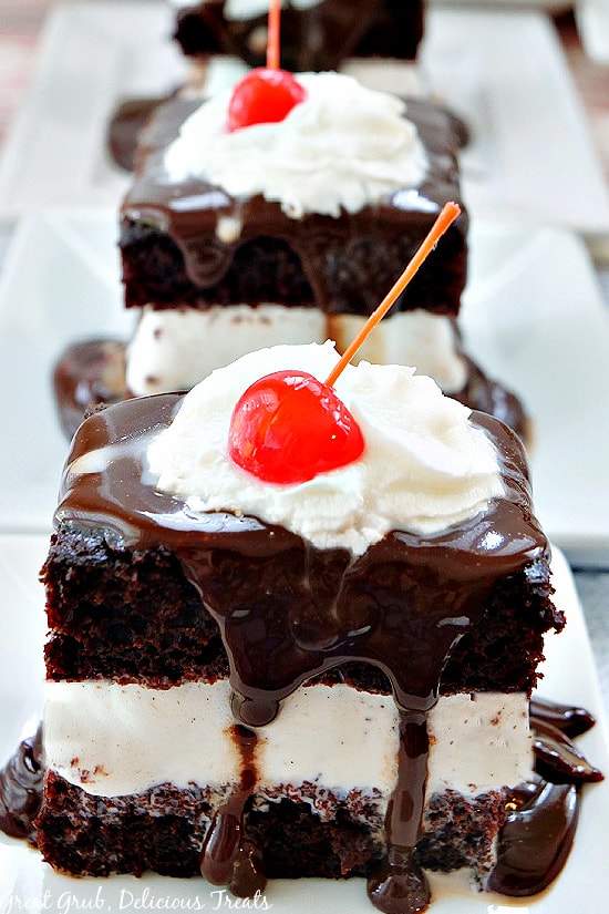 Three white plates lined up, all with a square piece of hot fudge cake on them, topped with hot fudge, whipped cream, and a cherry.