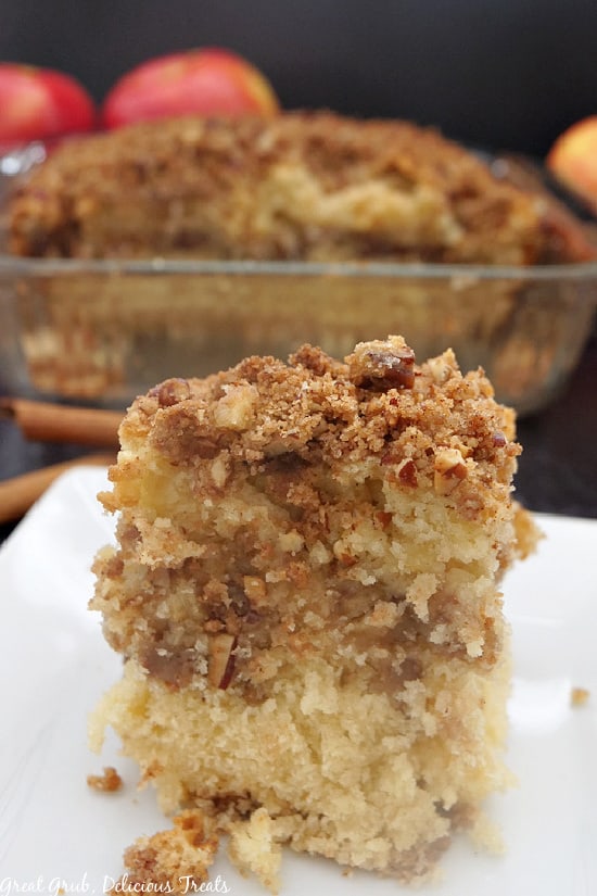 A piece of apple pecan coffee cake on a white plate, with the dish of coffee cake in the background.