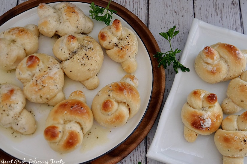 Garlic knots on a white round plate and a square white plate sitting on a white wood board.