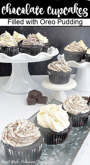 Chocolate cupcakes on a white cake stand with other cupcakes lined up on a silver plate.