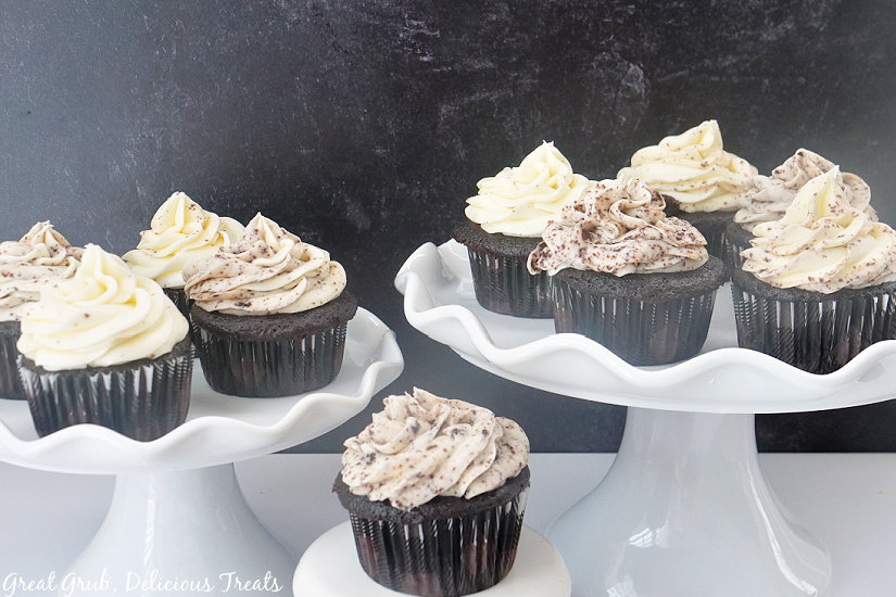 Two white cake stands with four chocolate cupcakes sitting on top, all topped with buttercream frosting and sprinkled with oreo crumbs.