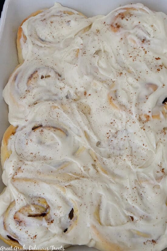 A white baker fill with freshly baked pumpkin spice cinnamon rolls and frosted with maple cream cheese frosting.