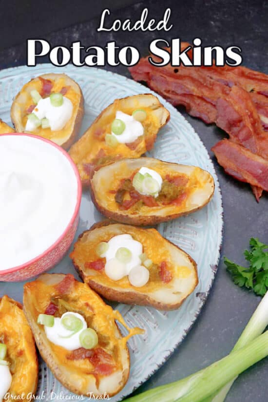A large plate lined with loaded potato skins, bacon in the background, and a small bowl of sour cream.