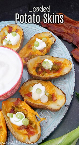 A large white plate with a small red bowl full of sour cream in the middle of the plate and loaded potato skins lined around the edges.