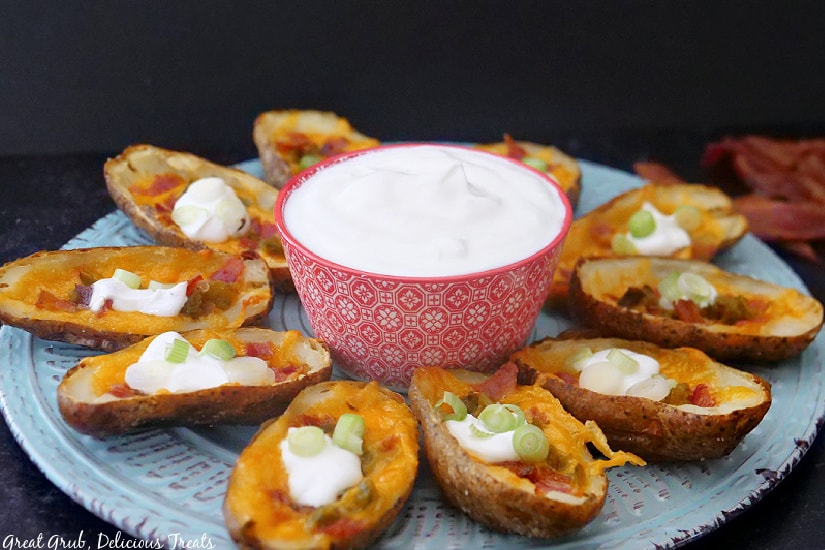 A large white plate with loaded potato skins around the edges and a small red bowl of sour cream in the middle of the plate.