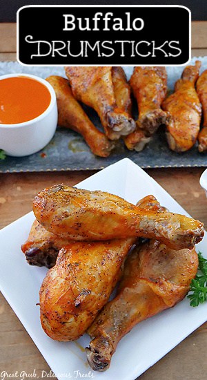 A white plate with four chicken drumsticks on in and a sliver plate in the background with more chicken legs and a small white bowl with buffalo sauce in it.