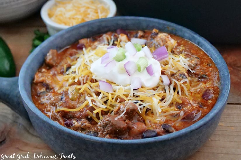 A close up pic of chili in a bowl topped with shredded cheese, sour cream, red onions and green onions.