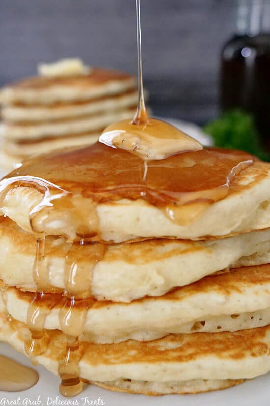 A white plate with a stack of four pancakes on it with a pad of butter on the top with syrup drizzling down.