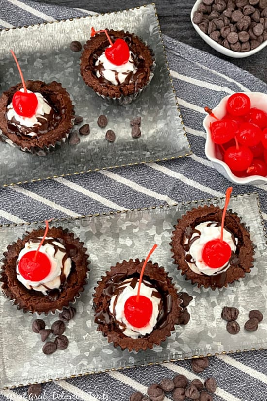 An overhead photo of a silver tray with three brownie cups on them with a white bowl of cherries in the background and chocolate chips laying around the plate.