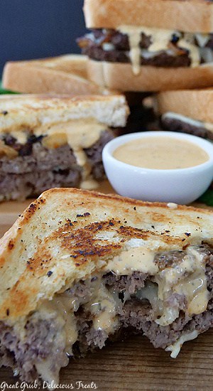 Half of a patty melt with a bite taken out of it on a wooden board with a small white bowl filled with a creamy sauce, with more patty melts in the background.
