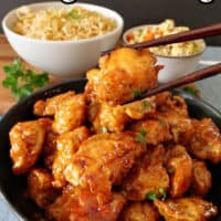 A black bowl with orange chicken in it with chopsticks holding a piece of chicken.
