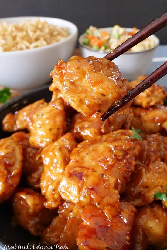 A close up of a piece of orange chicken held with chopsticks above a serving bowl.