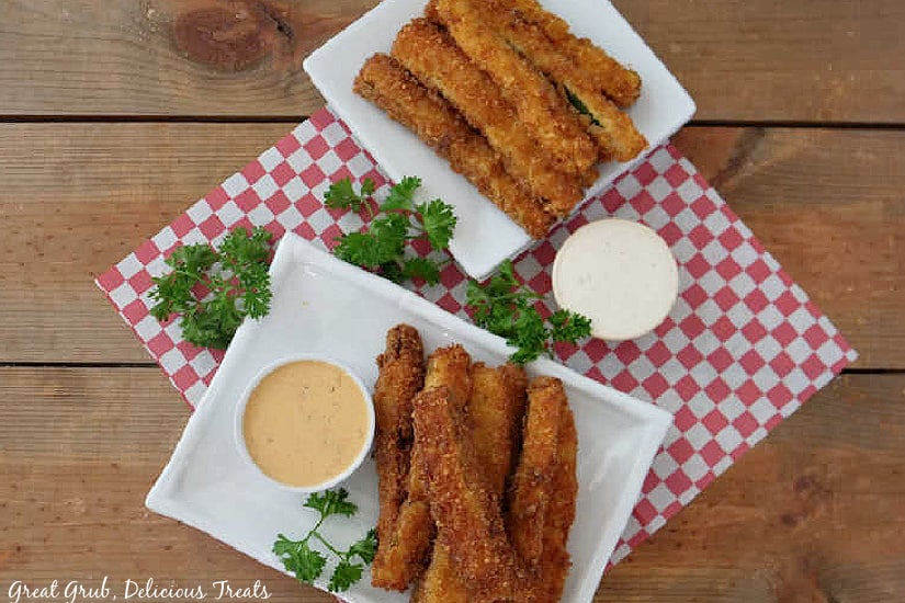 An overhead shot of two white plates sitting on a red and white placemat with fried zucchini strips on it with dipping sauce.