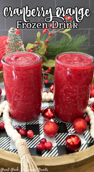 Two glasses filled with frozen cranberry orange and agave drink placed on a black and white round tray garnished with red holiday ornaments and white beads.