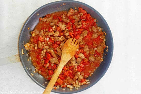 A skillet with cooked sausage, diced tomatoes, onions, garlic and seasoning in it with a wooden spoon in the skillet.
