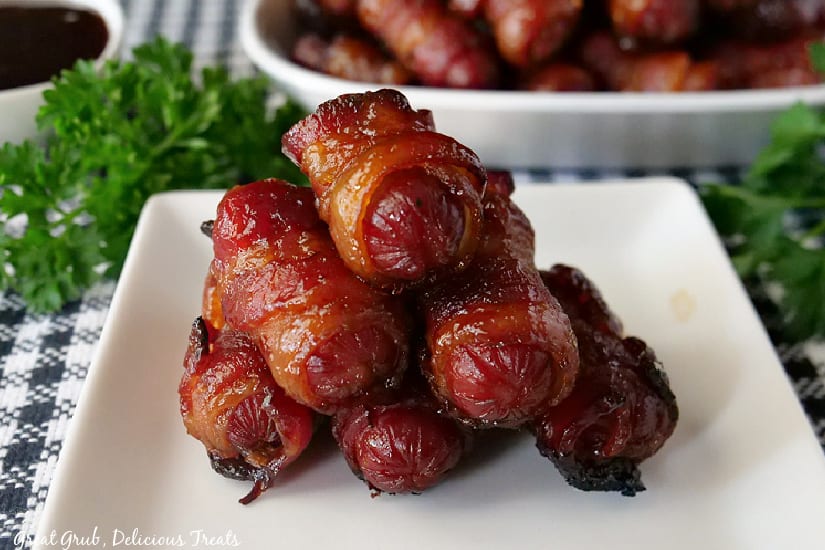 A black and white placemat with a small white plate with bacon wrapped little smokies on it with another bowl in the background with more smokies in it.