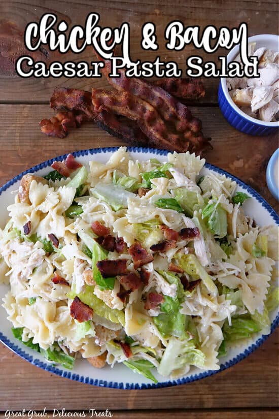 A wood surface with a white bowl with blue trim with a chicken Caesar pasta salad in it with slices of crispy bacon on the side.