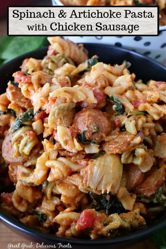 A close up of a black bowl filled with spinach and artichoke pasta with chicken sausage.