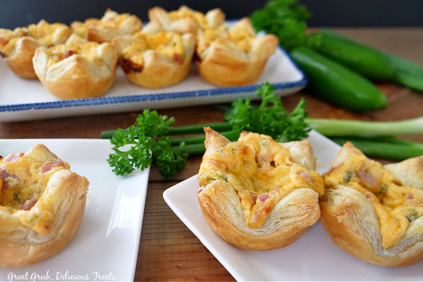 Three white square plates with ham and cheese pastries sitting on them, green onions, parsley, and jalapenos are in the back ground.