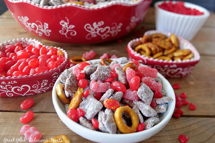 A white bowl with valentine muddy buddy mix in it with two red heart bowls filled with pretzels and red hot candies.