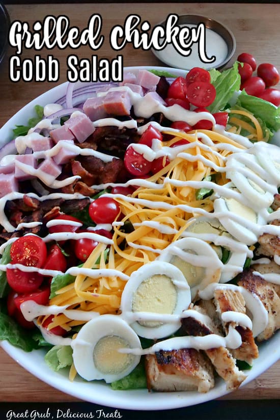 A large white bowl filled with lettuce, diced chicken and ham, bacon, sliced boiled eggs, grape tomatoes, cheddar cheese, and drizzled with ranch dressing. 