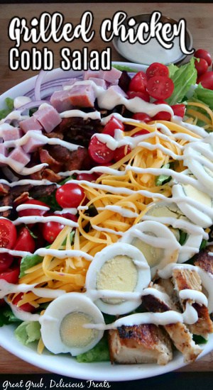 A large white bowl with lettuce, lined with sliced boiled eggs, grilled chicken, cheese, tomatoes, bacon, diced ham, drizzled with ranch dressing. 