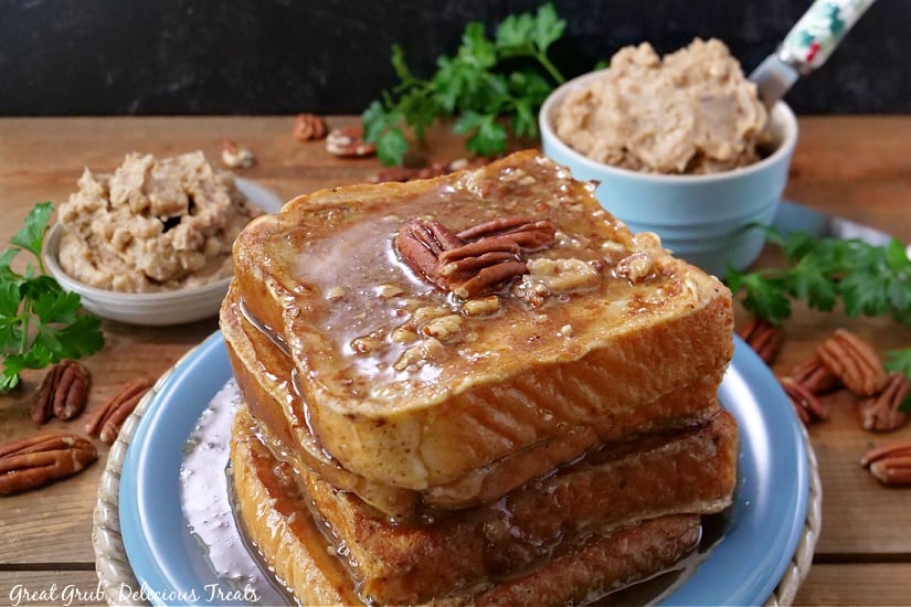 Four pieces of French toast stacked up on a blue plate with syrup and pecans on top, and two small bowls of whipped butter in the background.