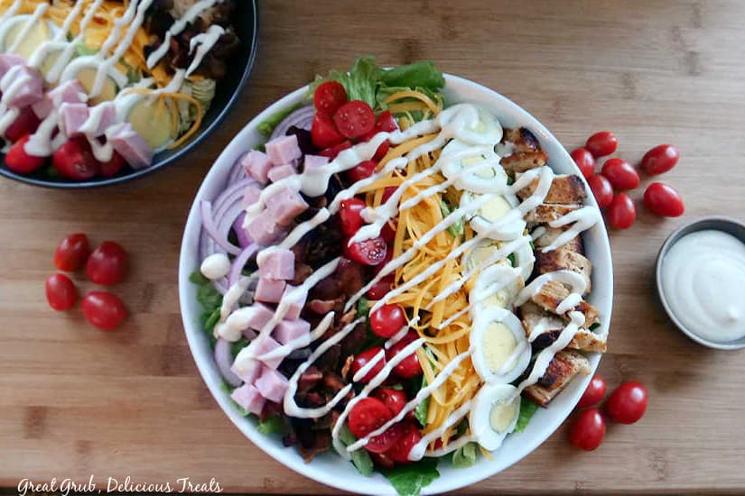 An overhead photo of a white bowl filled with salad, a black bowl in the background, and grape tomatoes laying around the plates of food. 