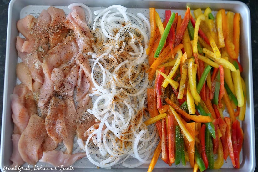 A large sheet pan with chicken, onions, and bell peppers on it, all topped with seasonings.
