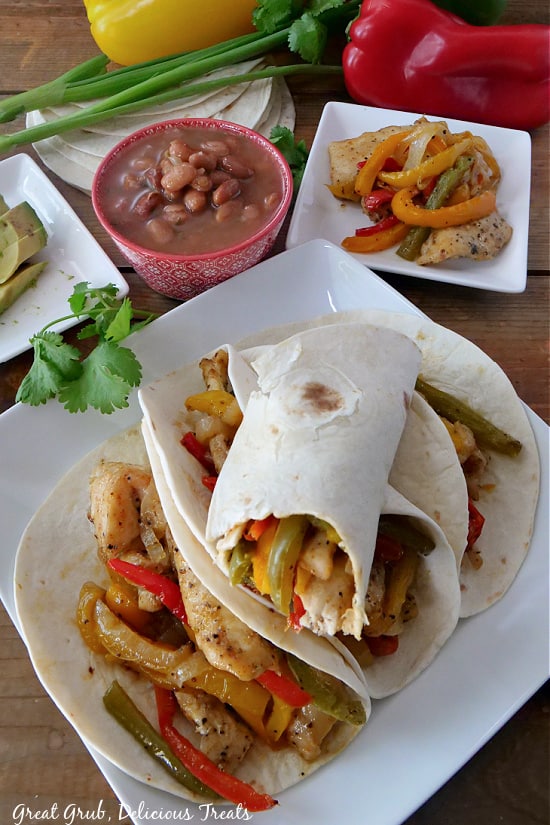 An overhead photo of chicken fajitas on a white plate, with a plate of chicken and vegetables in the background, and a small red bowl full of charro beans.