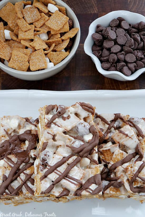 A white plate with three marshmallow treats on it with two white bowls filled with cereal and one with chocolate chips.