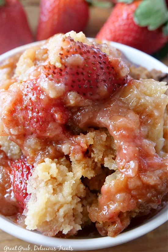 A close up picture of a white bowl of strawberry rhubarb cobbler with fresh strawberries in it.