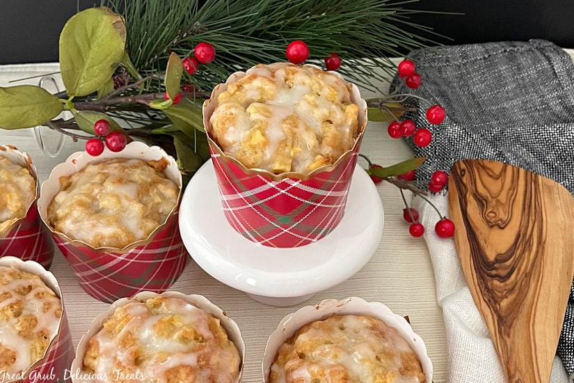 Apple muffins in red plaid cupcake liners, all topped with a glaze, with one muffin sitting on a white cupcake stand.