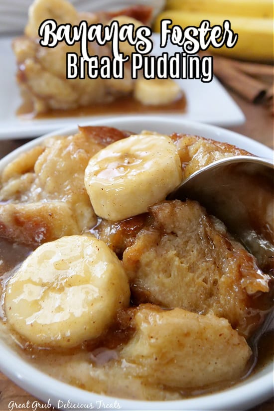 A white bowl filled with bananas foster bread pudding and a white plate in the background, along with cinnamon sticks and a banana in the back for decoration.