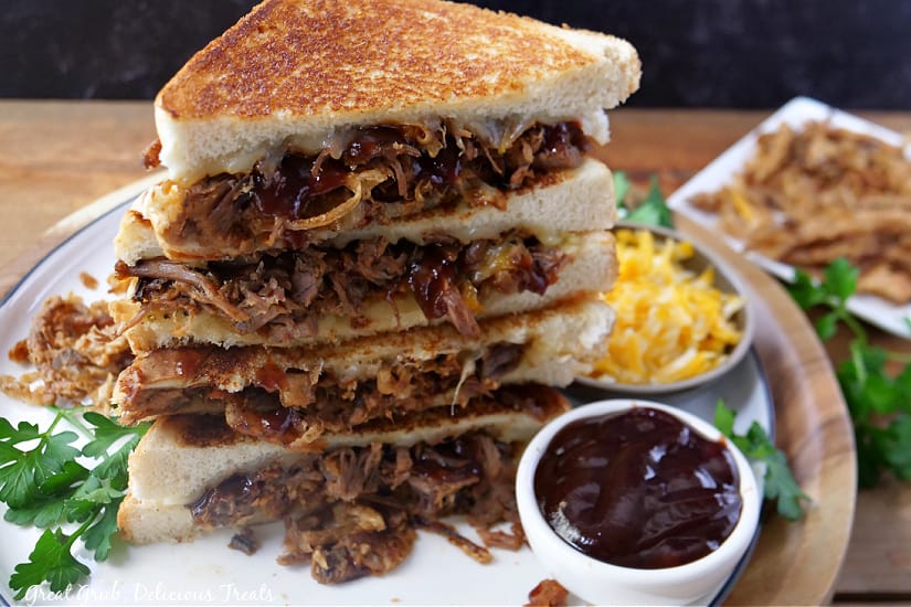 Two brisket grilled cheese sandwiches cut in half and stacked on top of each other with a bowl of shredded cheese and a bowl of barbeque sauce in the background.