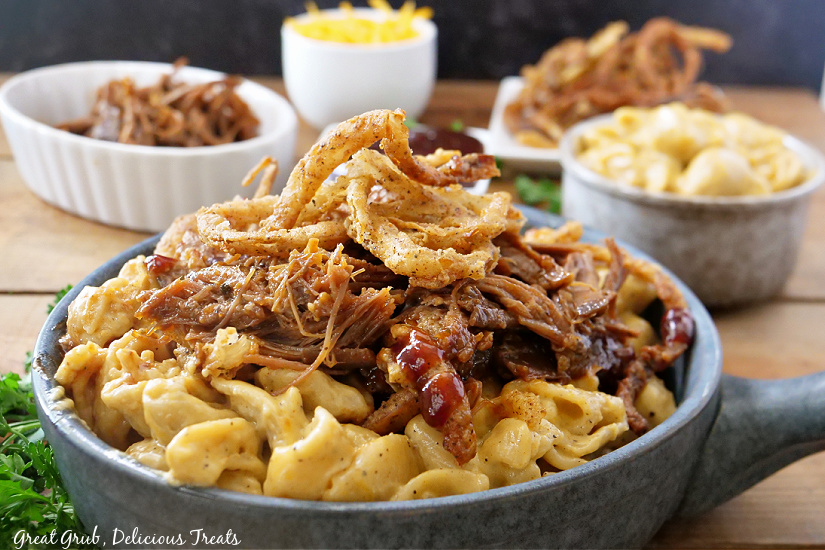 A large bowl of mac and cheese topped with brisket and onion strings, small bowls in the background filled with brisket, mac and cheese, shredded cheese, and a plate of onion strings.