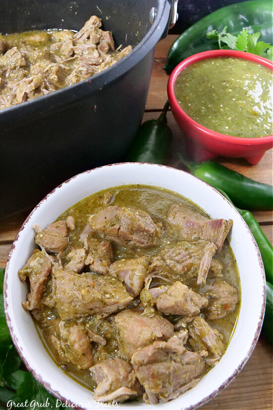 Chili verde recipe in a medium sized bowl, a small red bowl filled with salsa verde sauce in the background, sitting next to a Dutch oven.